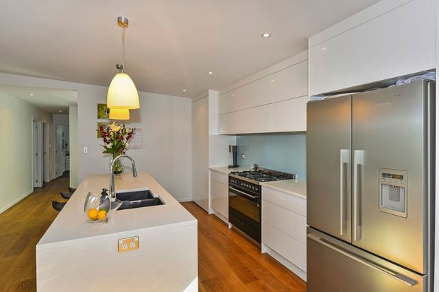 Bright contemporary kitchen with a large pale stone island, integrated sink and tap, white handleless cabinetry, stainless-steel refrigerator and freestanding oven with gas cooktop. Warm timber flooring, recessed ceiling lights and pendant lighting create a clean, inviting fit-out, with a hallway visible beyond.