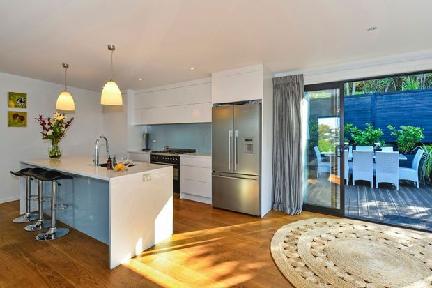 Bright contemporary open-plan kitchen with a white island, integrated white cabinetry, stainless-steel refrigerator and appliances, timber flooring, pendant lights, and a large glazed opening to a furnished outdoor deck. A round woven rug and dining setting are visible in the adjoining living and outdoor areas.