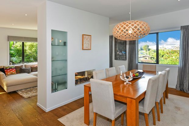Bright open-plan dining area with a large timber dining table and upholstered chairs, adjoining a living room visible to the left. The room features white walls, warm timber flooring, full-height grey curtains, large windows overlooking greenery, a built-in glass display niche, and a woven pendant light.