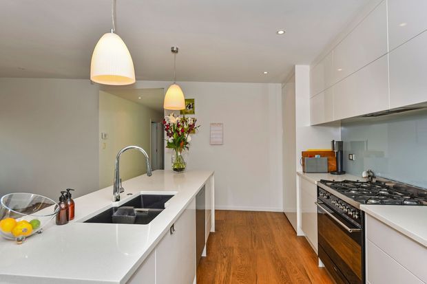 Bright contemporary galley-style kitchen with a large white island and integrated dark double sink, white cabinetry, pale glass splashback, stainless steel and black range cooker, timber flooring, and pendant lighting. A small dining area and floral arrangement are visible beyond the island.