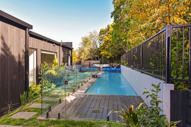 Exterior view of a narrow landscaped residential pool area, with a long rectangular swimming pool bordered by dark timber decking, frameless glass pool fencing, and a black metal privacy fence. A dark timber-clad building runs along the left side, while mature planting surrounds the space.