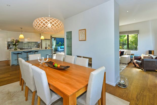 Bright open-plan dining area with a large timber dining table and upholstered dining chairs, opening directly to a modern kitchen with island and pendant lighting. A living room is visible beyond, with white walls, timber flooring, and generous natural light.