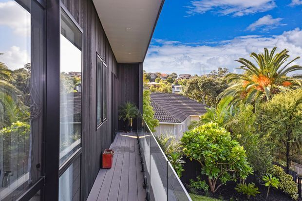 A narrow elevated balcony or terrace running along the side of a contemporary dark-clad house, with full-height glazing, timber-look wall cladding, a slim balustrade, and views over landscaped suburban gardens and neighbouring rooftops. Lush palms and planting create a bright, leafy outdoor setting beneath a blue sky.