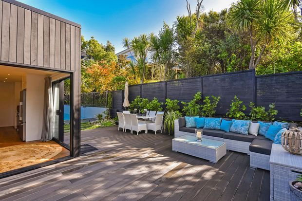 A sunny outdoor entertaining area with a spacious timber deck, built-in dark privacy fencing, an outdoor dining setting, and a large wicker sectional lounge with blue cushions. The deck connects directly to a contemporary glazed living space, with lush subtropical planting surrounding the private courtyard.