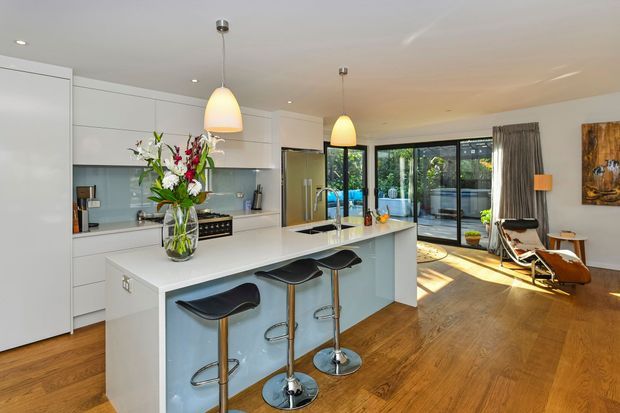 A bright, open-plan contemporary kitchen with white full-height cabinetry, a large white island with integrated seating, pale blue-grey backsplash, stainless-steel appliances and warm timber flooring. Three sculptural black bar stools line the island. Pendant lights hang above the island, while folding or sliding glass doors open to an outdoor area. A relaxed lounge corner with a leather chaise and side table is visible beyond the kitchen.