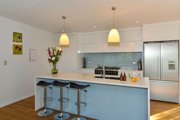 Bright contemporary kitchen with a large pale blue island, white cabinetry, integrated stainless-steel refrigerator, glossy light blue splashback, timber flooring, and three black bar stools. Two warm pendant lights hang above the island, while floral arrangements and small decorative items add colour.