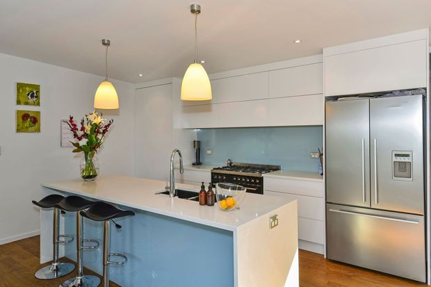Bright contemporary kitchen with a large white island featuring a pale blue side panel, integrated sink and curved chrome tap, three black bar stools, white handleless cabinetry, pale blue glass splashback, stainless-steel refrigerator and freestanding black range. Warm timber flooring and pendant lights add contrast.