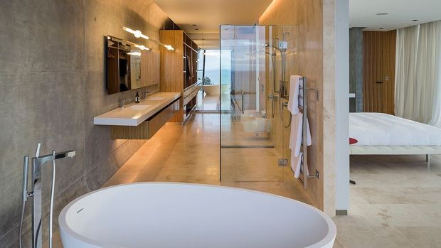 Luxurious contemporary bathroom with a freestanding oval bathtub in the foreground, a glass-enclosed shower, wall-mounted double vanity, warm timber cabinetry, and large-format stone-look wall and floor finishes. The bathroom opens directly to a bedroom visible on the right, with a bed and bedside furniture.
