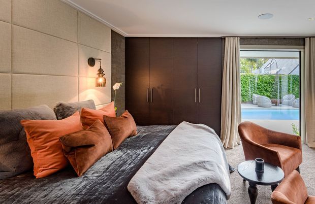 Warm, contemporary bedroom with a large upholstered bed, layered rust and brown cushions, dark wood built-in wardrobes, beige padded wall panels, and a small round side table with leather lounge chairs. Full-height curtains open to a private outdoor pool area visible through the glazing.