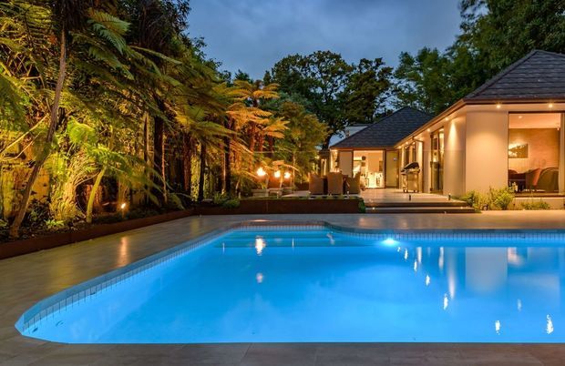 Twilight exterior view of a landscaped residential property with a large illuminated swimming pool in the foreground. A low-rise home with dark pitched roofs, warm cream rendered walls, broad glazed openings, and an outdoor entertaining area sits behind the pool, bordered by dense fern-filled planting and landscape lighting.