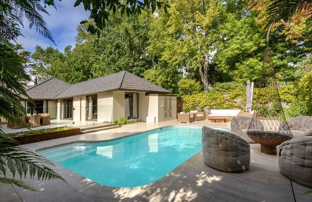 A landscaped outdoor pool area beside a low-slung pavilion with a hipped roof and extensive glass doors. The scene includes a turquoise swimming pool, paved terrace, outdoor seating, and dense tropical greenery, creating a private resort-like courtyard setting.
