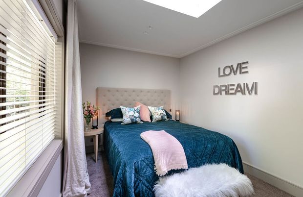 A bright, cozy bedroom with a large upholstered bed dressed in deep teal bedding, decorative pillows, and a pink throw. A wide window with white plantation blinds and curtains occupies the left wall, while a skylight provides additional natural light. The room has pale walls, carpeted flooring, a small bedside table, and decorative “LOVE DREAM” wall lettering.