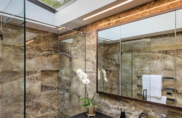 Luxurious contemporary bathroom featuring a large frameless glass shower enclosure, richly veined brown marble-look wall tiling, a dark stone vanity benchtop, and a wide framed mirror. A skylight and integrated linear ceiling lighting provide bright illumination, while an orchid adds a decorative touch.