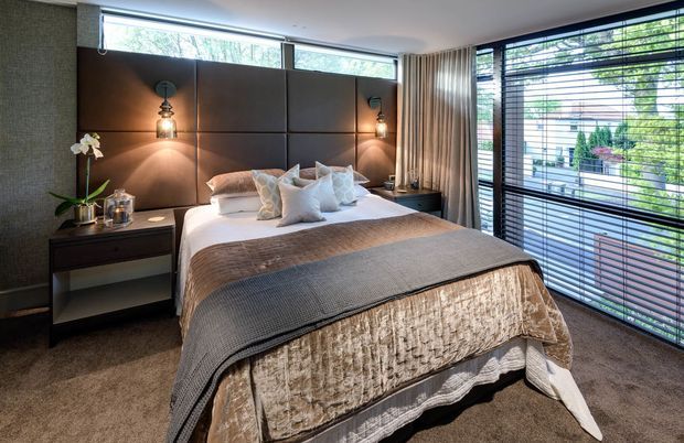 A sophisticated contemporary bedroom with a centrally positioned bed, layered neutral bedding, a large padded dark feature headboard, integrated wall-mounted bedside lighting, and compact dark bedside furniture. Full-height windows with horizontal blinds and draped curtains provide abundant natural light, while soft carpeting and textured fabrics create a warm, luxurious atmosphere.