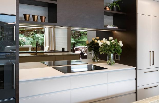 Contemporary kitchen with a large white island, integrated black induction cooktop, dark timber open shelving and cabinetry, tall white pantry cabinetry, and reflective splashback or mirrored wall panels. Natural light enters from the adjoining living area, with indoor plants and floral arrangements adding organic detail.
