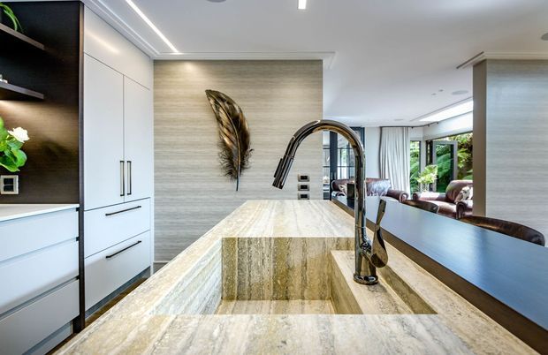 Contemporary open-plan kitchen viewed from the island, featuring a large waterfall-edge stone island with integrated double sink and dark sculptural tapware. White handleless cabinetry is paired with dark timber open shelving, while a textured light stone feature wall and recessed linear lighting add warmth and refinement. The kitchen opens toward a bright living area with extensive glazing and greenery visible beyond.