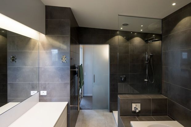 Contemporary bathroom with large dark grey tiled walls, a frameless glass walk-in shower, built-in tiled bench, wall-mounted shower fittings, and a white vanity with a large mirror. The space has recessed lighting and a restrained, high-contrast neutral palette.