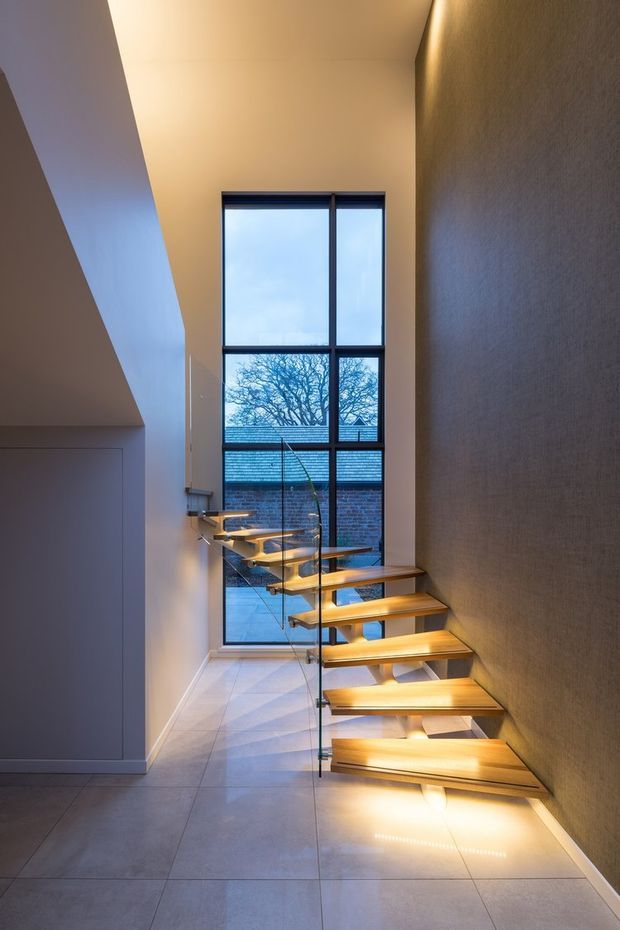 Interior view of a contemporary stairway with floating light-wood treads, concealed under-step lighting, and clear glass balustrades. The stair rises beside a tall floor-to-ceiling black-framed window overlooking a brick wall and tree, while large-format pale floor tiles and textured neutral walls create a refined modern setting.