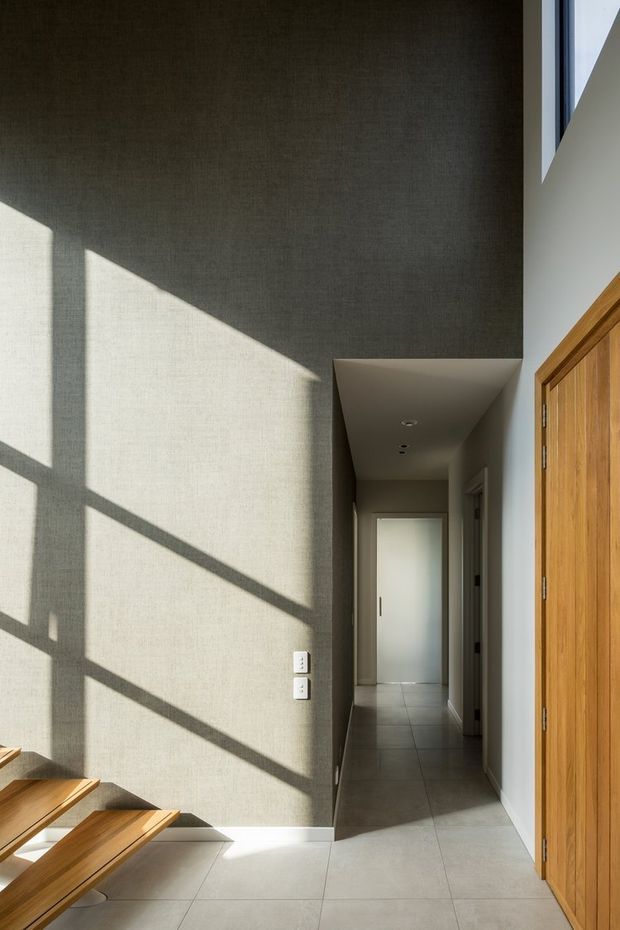 A narrow, sunlit interior hallway or entry passage with textured grey-beige walls, large pale floor tiles, recessed ceiling lights, and strong geometric shadows cast by glazing. Warm timber doors and a timber stair or bench edge add contrast, while a frosted glass door terminates the corridor.