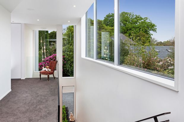 Bright, minimalist residential hallway or landing with white walls, recessed ceiling lights, grey carpet, and extensive floor-to-ceiling glazing overlooking leafy suburban gardens and neighbouring rooftops. A decorative chair and plants are visible at the end of the corridor, with a stair handrail partially visible in the foreground.
