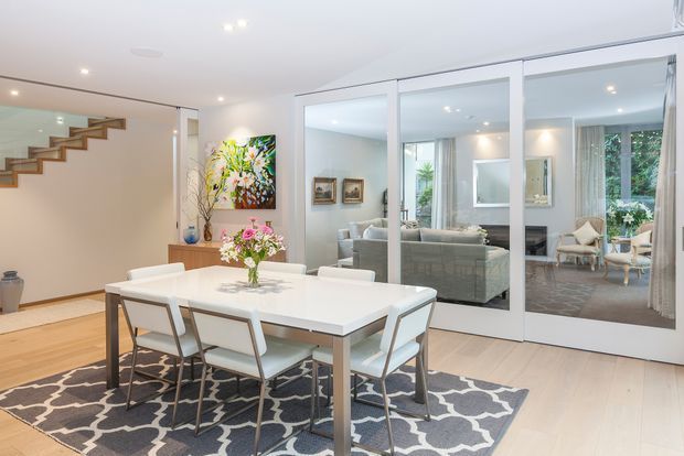 Bright, open-plan dining area with a white rectangular dining table and six upholstered chairs on a patterned rug. Light timber flooring, white walls, recessed ceiling lighting, contemporary artwork and floral styling create a fresh, refined interior. A floating timber staircase is visible at left, while large white-framed glazed doors provide a view and reflection into an adjoining living room with sofas and armchairs.