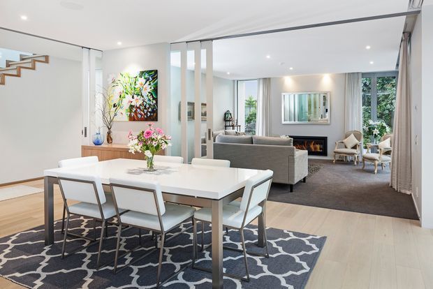 Bright open-plan interior combining a dining area and living room. A white rectangular dining table with contemporary chairs sits on a patterned rug in the foreground, while a grey sofa, fireplace, armchairs, artwork and large windows define the living area beyond. Pale timber flooring, white walls, recessed lighting and slim internal partitions create an airy, refined contemporary setting.