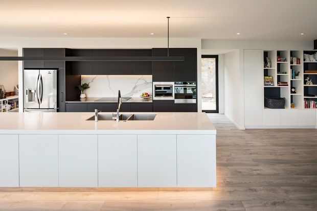 A bright, contemporary open-plan kitchen dominated by a large white island with integrated sink and tapware. The kitchen features dark charcoal cabinetry, a pale marble-look splashback, stainless-steel refrigerator and appliances, and a long linear pendant light. Timber-look flooring extends toward a white built-in shelving and storage wall, with a doorway and windows providing natural light.