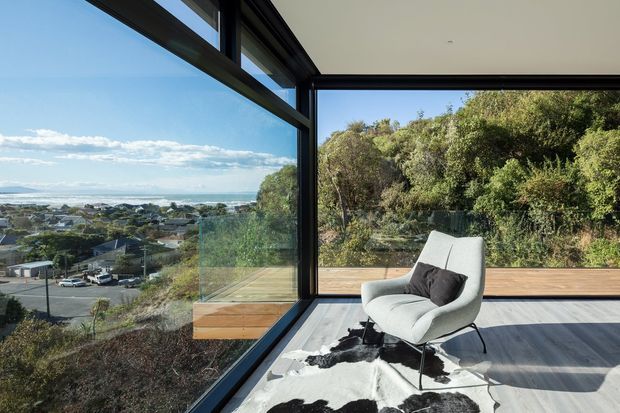 A bright, minimalist sitting area enclosed by floor-to-ceiling corner glazing, with a light upholstered lounge chair and dark cushion on a cowhide rug. Timber deck boards extend outside toward dense native vegetation, while the glazing frames a wide coastal suburban view.