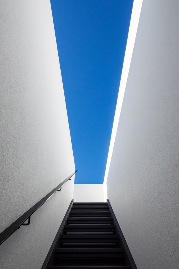 A narrow modern stairwell viewed upward, with dark timber or painted stair treads, white textured walls, a simple black handrail, and a striking blue ceiling or skylight opening above. The composition emphasizes the vertical geometry and strong contrast between the blue overhead plane, white walls, and dark staircase.