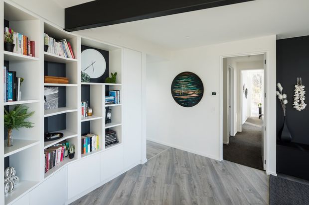 Bright contemporary hallway or living area with extensive built-in white shelving filled with books and decor, grey timber-look flooring, white walls, and a bold black ceiling beam and feature wall. A corridor leads to additional rooms, while wall art, plants, and sculptural accessories add visual interest.