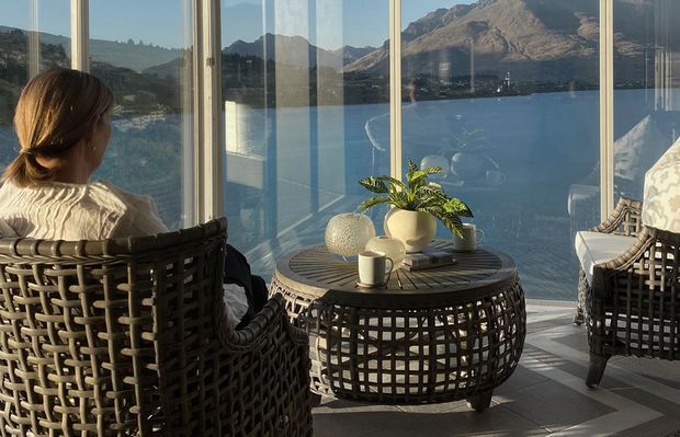 A tranquil sunroom or enclosed terrace with floor-to-ceiling glazing overlooking a deep blue lake and rugged mountains. Two woven lounge chairs face a round woven coffee table holding a cup, ceramic vessels, and a leafy plant. The space is warmly lit with a relaxed natural palette and strong indoor-outdoor connection.