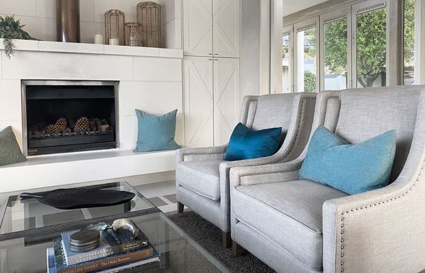 Bright, refined living room with a white built-in fireplace surround and cabinetry, two upholstered wingback armchairs, a glass coffee table, and blue accent cushions. Large windows bring in abundant natural light and greenery is visible outside.