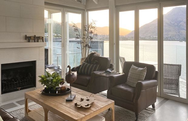 A refined living room with a built-in fireplace, timber coffee table, leather armchairs, and a large patterned rug. Floor-to-ceiling glazing and glazed doors open to a balcony or terrace with expansive waterfront and mountain views. The room is softly illuminated by warm natural light and features a neutral palette with timber and dark leather accents.