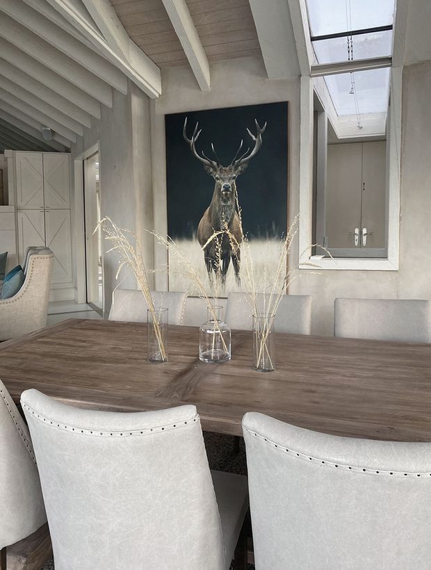 A bright, refined dining room with a long dark timber dining table, upholstered light dining chairs, a large deer artwork, pale textured walls, exposed white ceiling beams, and a skylit corridor or window beside built-in cabinetry. Dried grasses in glass vases provide a natural decorative accent.