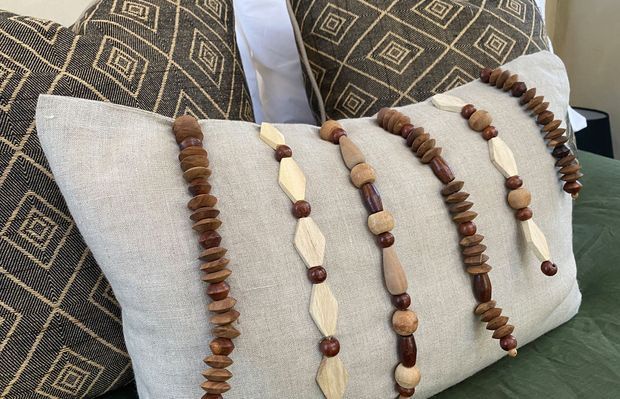 Close-up interior detail of a layered seating arrangement: a large natural linen cushion rests against dark upholstered cushions with repeating geometric diamond patterns. Several strands of wooden bead decoration are draped across the front cushion, creating a warm, textured, earthy accent.