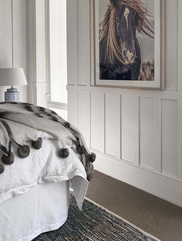 A softly styled bedroom corner featuring a white bed layered with a grey throw and tasselled pom-pom detailing, a patterned bedside lamp, white wall panelling, a neutral carpet, and a framed horse portrait. The room has a calm, monochromatic and traditional panelled aesthetic.