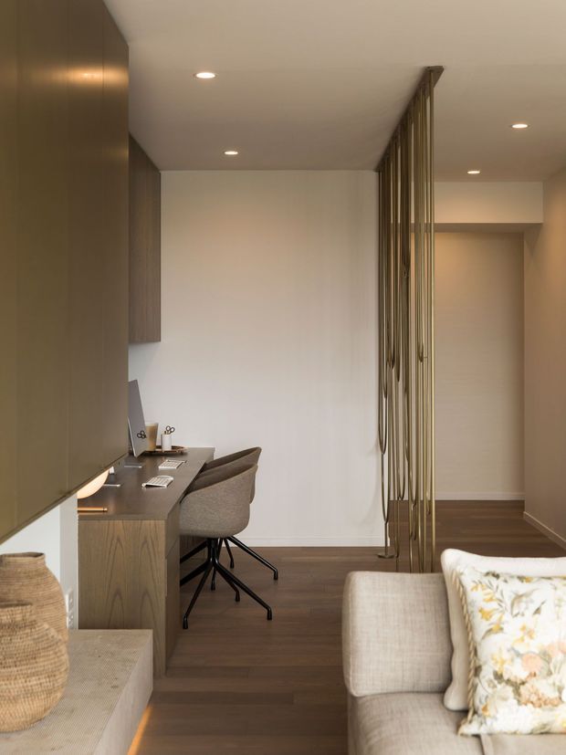 A warm, refined open-plan interior combining a compact home office with a bedroom or living area. A built-in timber desk with two upholstered swivel chairs sits beneath dark cabinetry, while a vertical brass-toned slatted divider separates the workspace from a partially visible upholstered bed or sofa. Dark timber flooring, softly lit white walls, recessed ceiling lights, and neutral textured furnishings create a calm, contemporary atmosphere.
