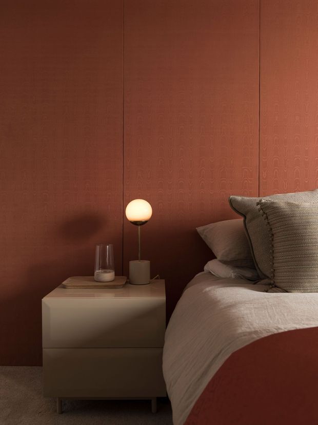 Warm, moody bedroom vignette featuring a bed with layered neutral bedding, a sculptural globe bedside lamp, and a minimalist floating bedside table. The room is defined by rich rust-brown patterned wall panelling and soft, low-level lighting.