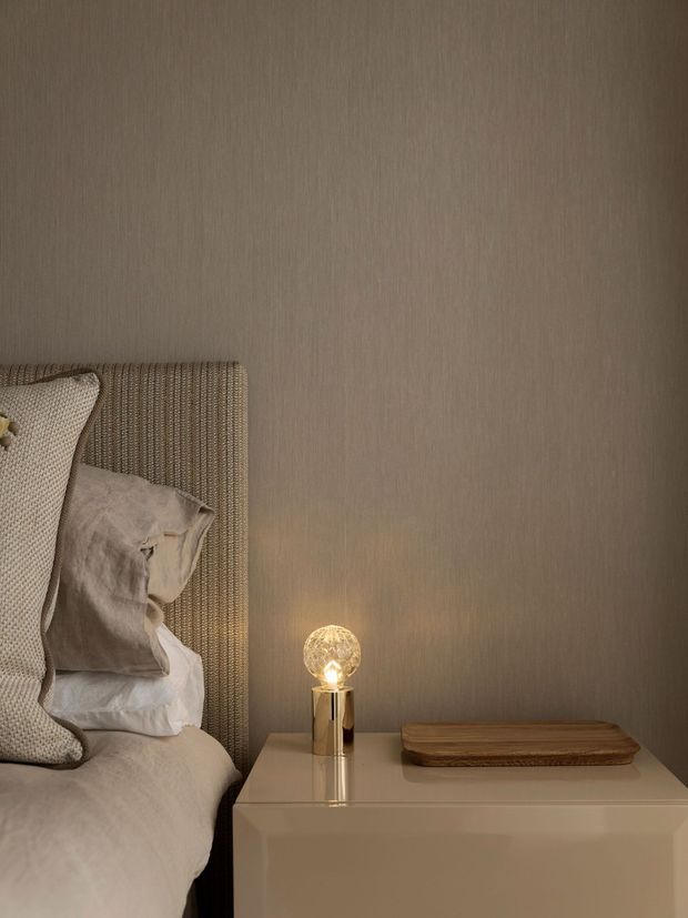 A close-up of a softly styled bedroom corner featuring a textured upholstered bedhead, layered neutral linen bedding and cushions, and a glossy cream bedside table. A small warm-glowing spherical lamp and wooden tray sit on the nightstand against a taupe textured wall, creating an intimate, refined atmosphere.