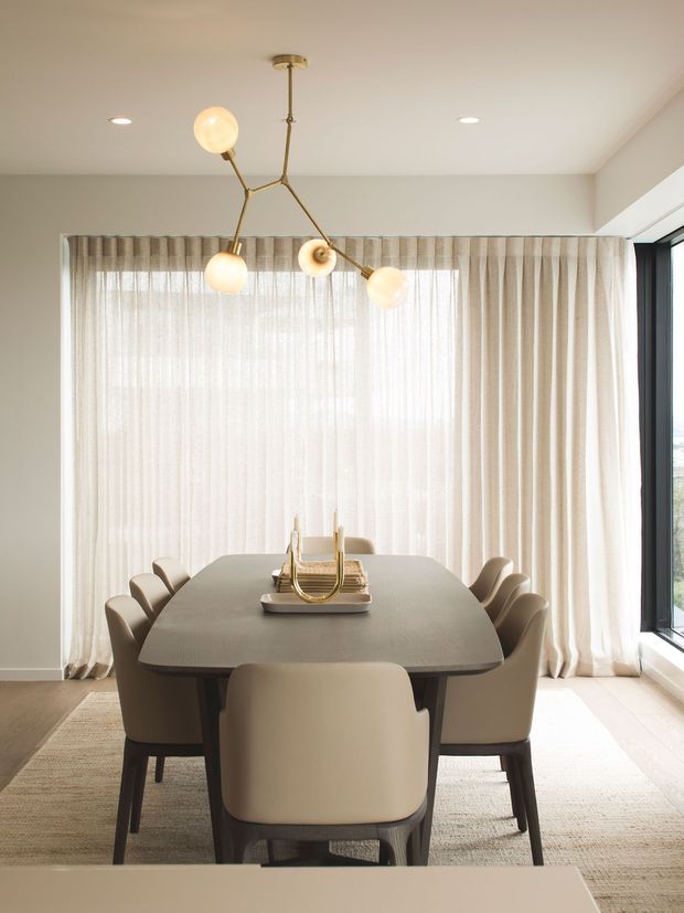 A refined contemporary dining room with a large rounded-rectangle dining table surrounded by upholstered dining chairs. Sheer full-height curtains span a broad window, complemented by a dark-framed corner window, recessed ceiling lights, and a sculptural branching pendant with glowing globe shades. Neutral beige, cream, and taupe finishes create a soft, elegant atmosphere.