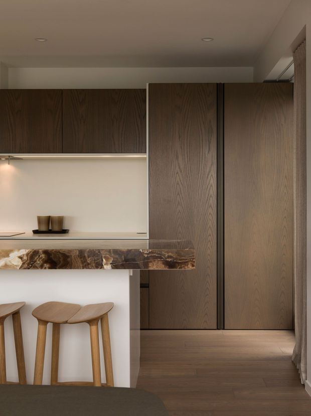 A refined open-plan interior featuring dark timber cabinetry and full-height wood-panelled storage, a compact stone-clad breakfast counter with sculptural bar stools, and integrated warm under-cabinet lighting. The space has a restrained, sophisticated palette of dark wood, cream walls, and richly veined brown stone, with a hallway or adjoining area visible beyond the joinery.
