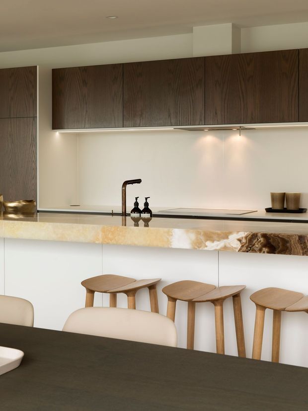 A refined contemporary kitchen with dark timber overhead cabinetry, a pale integrated backsplash, and a long stone island with a dramatic veined natural-stone edge. Warm timber stools line the island, while a dark dining table and upholstered chairs occupy the foreground. Under-cabinet lighting and restrained decor create a sophisticated, warm atmosphere.