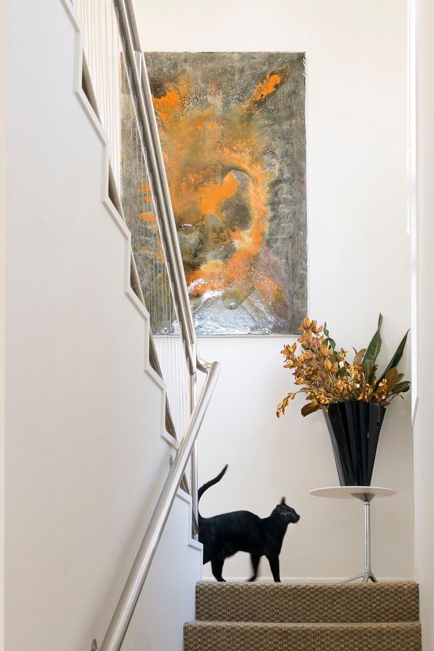 Bright, narrow residential stairway with carpeted steps, white walls, slim metal handrails and a geometric wall detail along the staircase. A large abstract artwork in grey, orange and metallic tones hangs at the landing beside a tall black vase with dried foliage. A black cat is visible on the stairs.