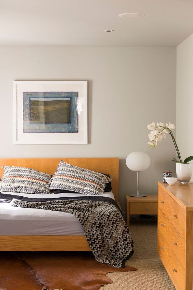 A warm, softly lit bedroom featuring a natural timber platform bed with a matching timber bedside table and dresser. The bed is dressed with black, white, and grey geometric-patterned cushions and throw, set against light greige walls. A round white table lamp, framed abstract artwork, and a potted white orchid complete the calm, contemporary interior.