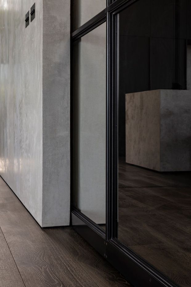 Close-up interior view of a dark, refined hallway or entry zone featuring wide dark timber flooring, a textured pale grey plaster wall, and a full-height black-framed mirrored or glazed partition. Through the opening, a dark room with a substantial stone or concrete island/cabinet is visible. The composition emphasizes strong vertical lines, black trim, natural material textures, and low-key lighting.