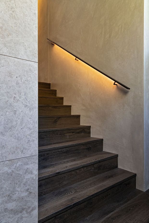 A contemporary interior stairway with broad dark timber treads rising along a textured warm beige plaster wall. A slim dark handrail incorporates warm linear LED lighting beneath it, while the adjacent wall is clad in large pale stone-look panels. The composition is minimalist, refined, and warmly illuminated.