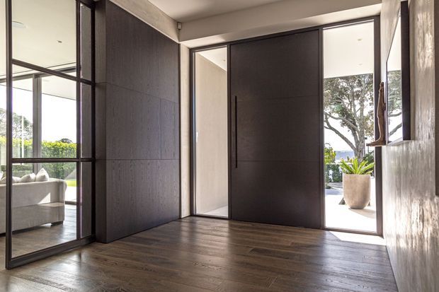 A refined residential entry foyer with a large dark pivot front door, full-height glazed sidelights, black-framed internal glazing, dark timber flooring, and textured dark wall panelling. A living room is visible through the glazed partition, while a bright landscaped exterior is visible through the open doorway. The space has a contemporary, warm, high-contrast character.