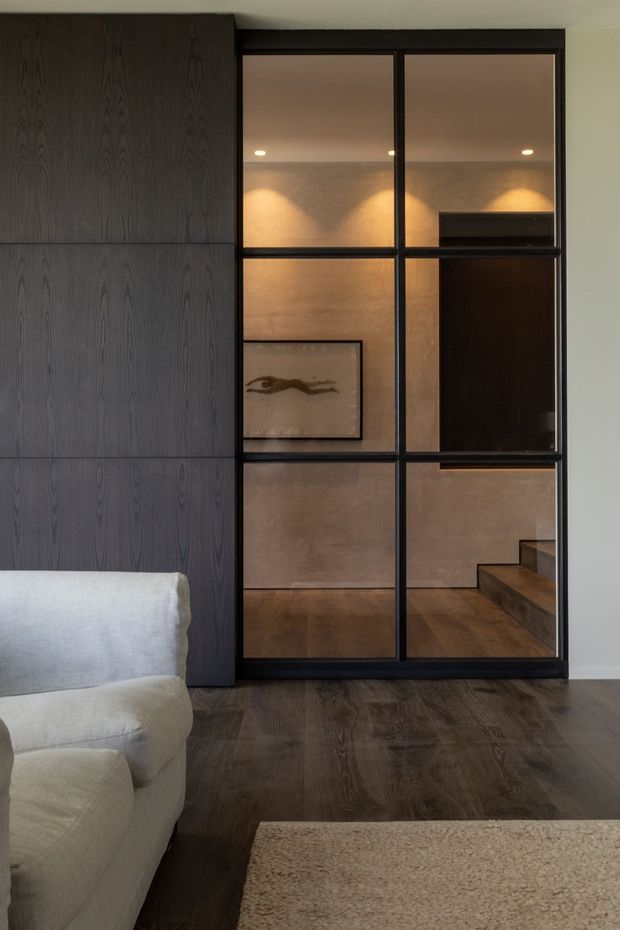 A refined living room with dark timber flooring and full-height dark wood wall panelling beside a black-framed internal glazed partition. The glazing reveals a warmly lit hallway or stair area with textured beige walls, timber flooring, artwork, and timber steps. A pale upholstered sofa and neutral textured rug occupy the foreground. The scheme is restrained, warm, and contemporary with strong dark-versus-light contrast.