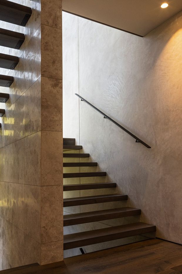 Interior view of a modern open-riser staircase. Dark timber treads project from a light textured plaster wall, with a slim dark metal handrail and a tall tiled or stone-clad central wall. Recessed ceiling lighting and concealed illumination beneath the treads create a warm, refined atmosphere.