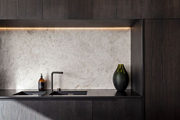 Close-up view of a sophisticated dark timber kitchen or bar-style joinery installation. A pale textured stone backsplash is illuminated by concealed warm LED strip lighting beneath overhead cabinetry. The dark stone benchtop includes an undermount sink and slim matte-black tap, with dark timber cabinetry and a sculptural black vase completing the refined, moody composition.